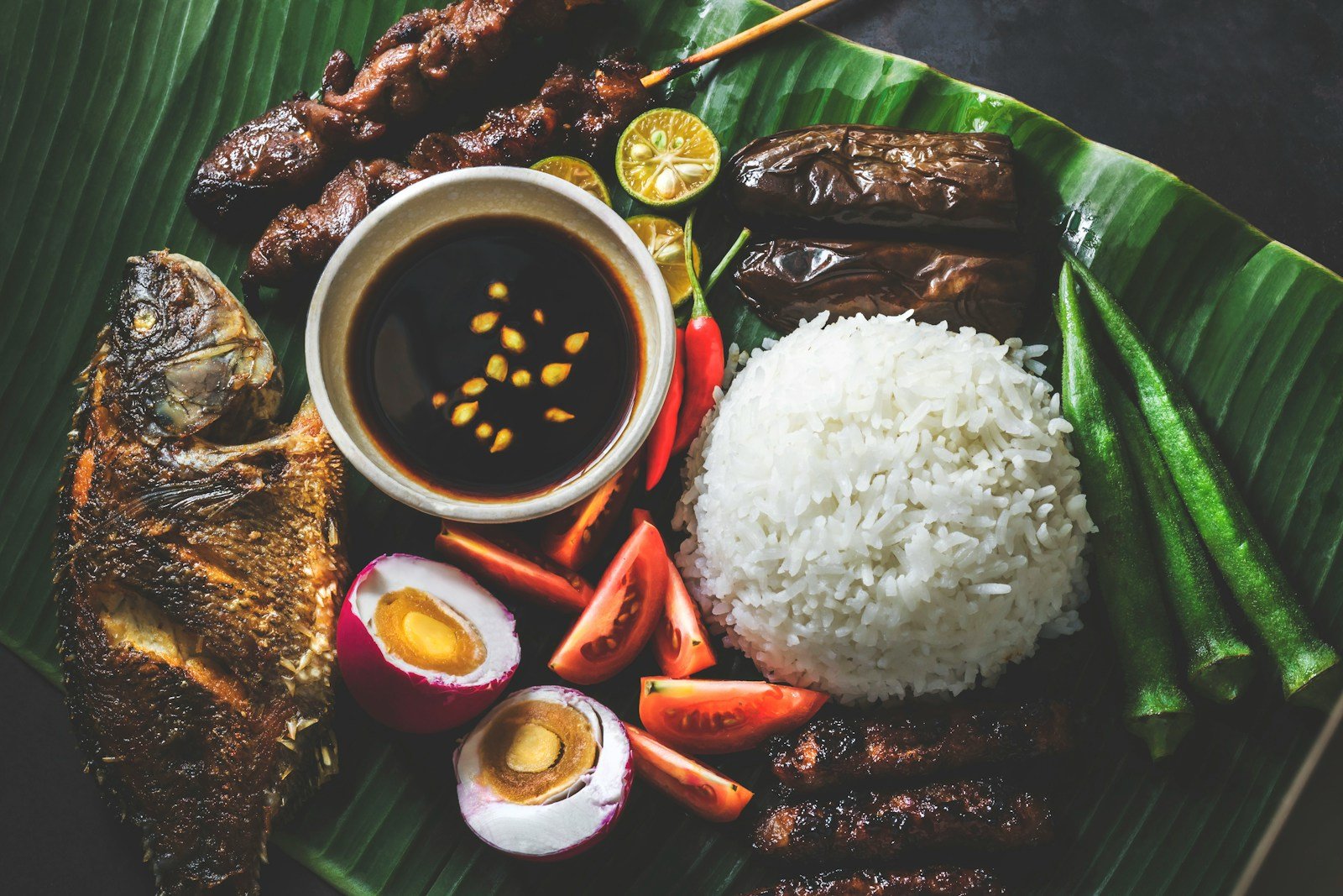 A Filipino meal laid out on a banana leaf: grilled fish, rice, salted eggs, tomatoes, okra, eggplant and skewers around a bowl of dipping sauce